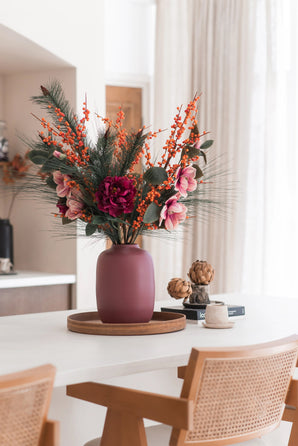 Het Zijden kunstbloemen boeket Cozy Winter in een mauve vaas, met roze bloemen, oranje bessen en dennentakken, siert een rond houten dienblad op een witte tafel voor een gezellige, moderne herfst/winterlook met houten stoelen en beige gordijnen.