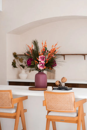 Een mauve vaas met het Zijden kunstbloemen boeket Cozy Winter - perfect voor herfst winterdecoratie - staat op een witte toonbank tussen houten stoelen, met boeken, artisjokken, planken en een witte pot met takken in de buurt.