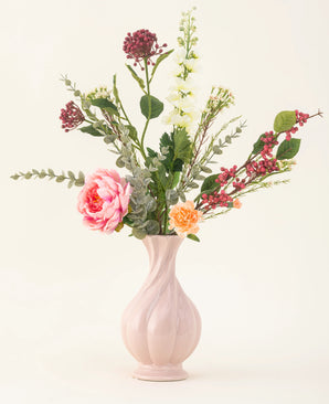 Le bouquet de fleurs artificielles en soie Pretty Petite, dans un vase rose pastel, contient des fleurs roses et orange faites à la main, des fleurs blanches, des feuilles vertes et des baies rouges - parfait pour un intérieur durable sur un fond clair uni.