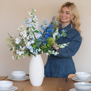 Een glimlachende vrouw in een blauwe spijkerjurk schikt het Zijden kunstbloemen boeket Cool Breeze in een gestructureerde witte vaas op een eettafel gedekt met wit servies en goud bestek.