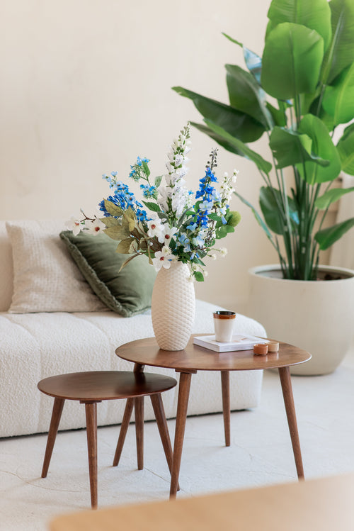 Dans un salon confortable se trouvent deux tables gigognes en bois avec un vase blanc contenant le bouquet de fleurs artificielles en soie Cool Breeze, ainsi qu'une tasse, un livre et une bougie. Un canapé blanc avec des coussins verts et une grande plante en pot complètent le décor aéré.