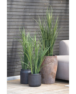 Une plante artificielle UV verte de gazon artificiel de 120 cm se dresse sur une terrasse en bois à côté d'un canapé gris clair, contre un mur horizontal sombre de planches de bois.