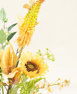 Le bouquet de fleurs artificielles en soie Amber Light comprend un tournesol jaune, une longue fleur épineuse jaune-orange, des feuilles vertes et de petites fleurs blanches sur un fond crème clair avec une lueur ambrée.