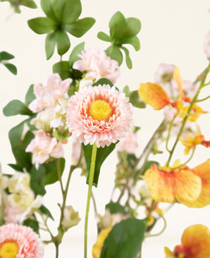 Un gros plan du bouquet de fleurs artificielles en soie Blossom Harmony, avec une fleur rose ressemblant à une marguerite avec un cœur jaune, entourée de fleurs rose clair et d'orchidées jaune orangé, sur un fond clair.