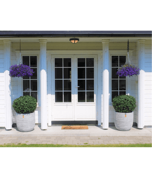 Une double porte blanche avec panneaux de verre est encadrée de deux Buis Artificiels sur tige UV d60 cm et de paniers suspendus à fleurs violettes, contre une maison blanche avec piliers et un petit tapis d'accueil à l'entrée.