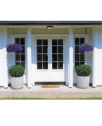 Une double porte blanche avec panneaux de verre est encadrée de deux Buis Artificiels sur tige UV d60 cm et de paniers suspendus à fleurs violettes, contre une maison blanche avec piliers et un petit tapis d'accueil à l'entrée.
