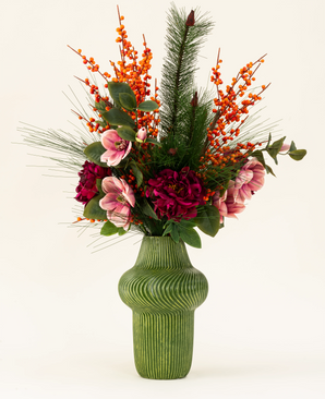 Het Zijden kunstbloemen boeket Cozy Winter presenteert een groene ribbelvaas met donkerrode en roze bloemen, oranje bessen, dennentakken en groene bladeren - perfect als herfst-winter decoratie tegen een lichte achtergrond.