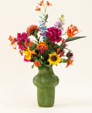 Un vase vert structuré montre le bouquet de fleurs artificielles en soie Colorful Spirit, avec des tournesols, des pivoines roses, des fleurs orange et violettes et une verdure luxuriante sur un fond clair uni.