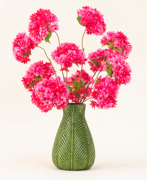 Un vase vert avec un motif de feuilles présente le bouquet de fleurs artificielles en soie Cherry Blossom Pink - des fleurs de cerisier en soie rose vif et moelleuses avec des feuilles vertes - sur un fond clair uni.