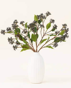 Un bouquet de fleurs artificielles en soie Baies japonaises Bleu Acier avec de longues tiges, des baies violet foncé et des feuilles d'acier se dresse dans un vase à texture blanche sur un fond clair uni.