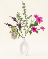 Het Zijden kunstbloemen boeket Lavender Skies, gerangschikt in een moderne witte vaas, bevat roze en paarse kunstbloemen, gele accenten en weelderig groen gebladerte - prachtig tegen een effen lichte achtergrond.