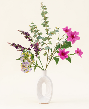 Het Zijden kunstbloemen boeket Lavender Skies, gerangschikt in een moderne witte vaas, bevat roze en paarse kunstbloemen, gele accenten en weelderig groen gebladerte - prachtig tegen een effen lichte achtergrond.