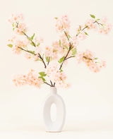 Un vase en céramique blanche avec une large ouverture ovale montre plusieurs bouquets de fleurs artificielles en soie à feuilles vertes, des branches rose clair de fleurs de cerisier sur un fond clair simple.