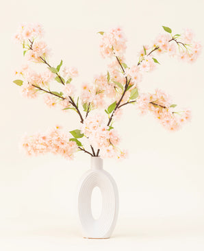 Un vase en céramique blanche avec une large ouverture ovale montre plusieurs bouquets de fleurs artificielles en soie à feuilles vertes, des branches rose clair de fleurs de cerisier sur un fond clair simple.