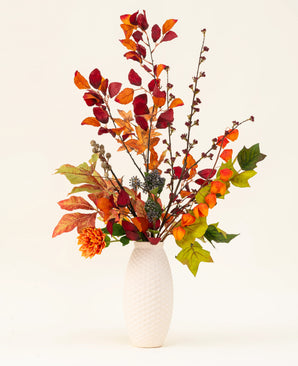 Een vaas met witte structuur toont het Zijden kunstbloemen boeket Chestnut Charm, met herfstbladeren, oranje bloemen en groen gebladerte - een elegant kunstarrangement tegen een effen lichte achtergrond.