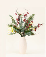Het Zijden kunstbloemen boeket Midwinter Dream heeft groene bladeren, dennentakken, rode bessen en witte bloemen in een vaas met witte structuur - perfect voor een sereen winterboeket tegen een effen lichte achtergrond.
