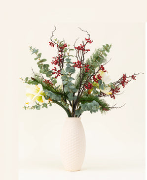 Het Zijden kunstbloemen boeket Midwinter Dream heeft groene bladeren, dennentakken, rode bessen en witte bloemen in een vaas met witte structuur - perfect voor een sereen winterboeket tegen een effen lichte achtergrond.