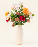 Het Zijden kunstbloemen boeket The Wild Bunch, met rode rozen, Real Touch Dahlia, roze bloemen en groene bladeren in een vaas met witte structuur, wordt getoond tegen een lichte achtergrond.