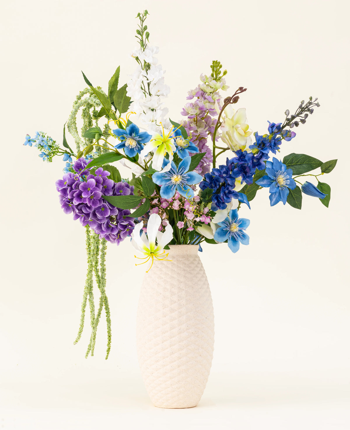 Un vase blanc structuré contient le bouquet de fleurs artificielles en soie Midnight Valley, composé de fleurs Real Touch bleues, violettes et jaunes vibrantes avec plusieurs feuilles vertes sur un fond clair.