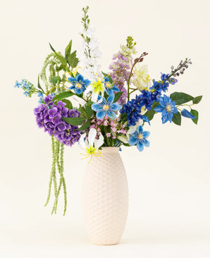 Een vaas met witte structuur bevat het Zijden kunstbloemen boeket Midnight Valley, met levendige blauwe, paarse en gele Real Touch bloemen met verschillende groene bladeren tegen een lichte achtergrond.