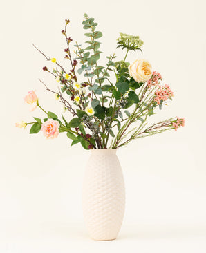 Een vaas met witte structuur toont het Zijden kunstbloemen boeket Blush Blossom, met echte bloemen - lichtroze rozen, gele bloemen en groen gebladerte - tegen een effen lichte achtergrond.