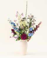 Un vase blanc avec le bouquet de fleurs artificielles en soie Purple Rain montre des fleurs artificielles violettes et blanches éclatantes et des feuilles vertes, un exemple de l'artisanat hollandais sur un fond clair.