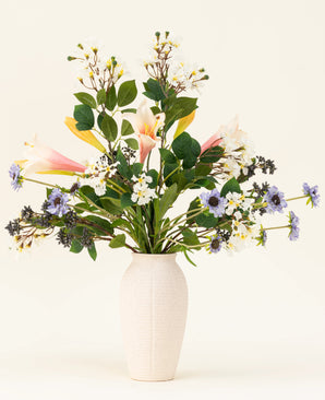 In een witte vaas staat het Zijden kunstbloemen boeket Blush & Bloom - met roze lelies, witte en paarse bloemen en weelderige groene bladeren - tegen een lichte achtergrond.