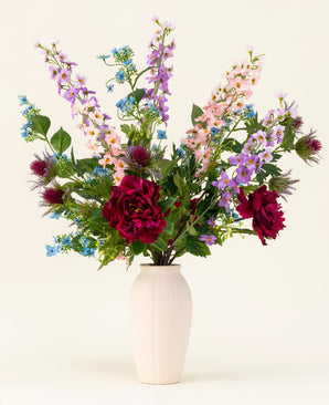 In een vaas met witte structuur staat het Zijden kunstbloemen boeket Moonlit Meadow - rode pioenrozen, roze en paarse riddersporen, blauwe bloemen, groene bladeren en distelachtige bloemen - tegen een effen, lichte achtergrond.