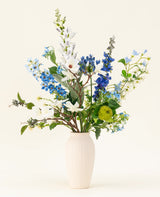 Het Zijden kunstbloemen boeket Cool Breeze, met witte lelies, blauwe ridderspoor en groen gebladerte in een crèmekleurige vaas, steekt af tegen een effen achtergrond voor een verfrissende touch.