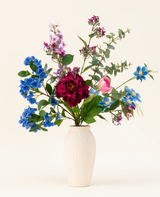 Een vaas met witte structuur toont het Zijden kunstbloemen boeket Painted Petals, met levendige blauwe, paarse, roze en rode zijden bloemen met groene bladeren en eucalyptustakken tegen een effen lichte achtergrond.
