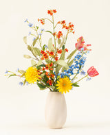 Het Zijden kunstbloemen boeket Lemonade Garden heeft echt getinte zonnebloemen, roze tulpen, blauwe riddersporen, oranje bloesems en groen gebladerte in een beige vaas tegen een effen lichte achtergrond.