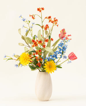 Het Zijden kunstbloemen boeket Lemonade Garden heeft echt getinte zonnebloemen, roze tulpen, blauwe riddersporen, oranje bloesems en groen gebladerte in een beige vaas tegen een effen lichte achtergrond.