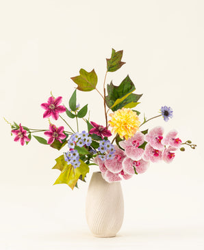 Het Zijden kunstbloemen boeket Aurora Garden heeft echte aanraakbloemen in roze, geel, blauw en paars met groene bladeren, gerangschikt in een witte keramieken vaas tegen een effen lichte achtergrond.