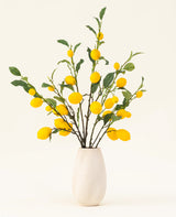 Un vase blanc texturé montre le bouquet de fleurs artificielles en soie Citrons, avec des feuilles vertes et des citrons jaunes vifs sur un fond clair.