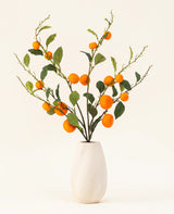 Le bouquet de fleurs artificielles en soie Mandarin a un vase texturé blanc et des branches de feuilles vertes et de mandarines artificielles orange vif, qui ajoutent une touche vivante sur un fond clair uni.