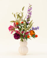 Un vase blanc et texturé présente le bouquet de fleurs artificielles en soie Colorful Svenski, composé de fleurs artificielles violettes, orange, roses et bleues vibrantes avec des feuilles vertes qui égayent n'importe quel espace sur un fond clair simple.