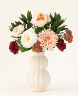 Een crèmekleurige vaas van keramiek met structuur toont het Zijden kunstbloemen boeket Jewel Bloom met roze, witte en donkerrode Real Touch bloemen en groene bladeren tegen een effen lichte achtergrond.