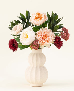 Een crèmekleurige vaas van keramiek met structuur toont het Zijden kunstbloemen boeket Jewel Bloom met roze, witte en donkerrode Real Touch bloemen en groene bladeren tegen een effen lichte achtergrond.