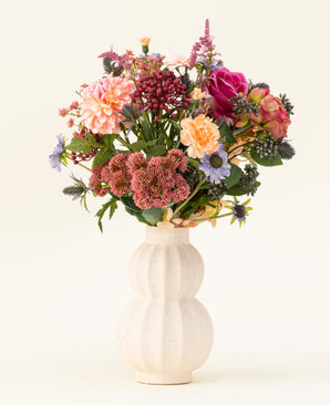 In een witte, gebogen vaas van keramiek staat het Zijden kunstbloemen boeket Pretty Pastel - een kleurrijke mix van duurzame bloemen zoals roze rozen, dahlia's, paarse en oranje bloemen en groen - prachtig gerangschikt tegen een lichte achtergrond.