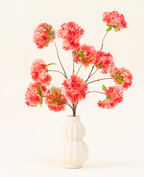Le bouquet de fleurs artificielles en soie, fleurs de cerisier et corail, présente des fleurs de cerisier artificielles rose vif et corail avec des feuilles vertes, élégamment disposées dans un vase blanc sculpté sur un fond clair.