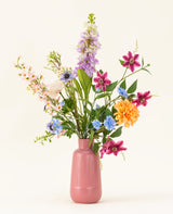 Het Zijden kunstbloemen boeket Bloom & Glow heeft roze, paarse, blauwe, oranje, witte en gele bloemen met groene bladeren in een roze vaas van keramiek tegen een lichte achtergrond.