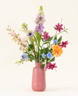 Het Zijden kunstbloemen boeket Bloom & Glow heeft roze, paarse, blauwe, oranje, witte en gele bloemen met groene bladeren in een roze vaas van keramiek tegen een lichte achtergrond.