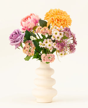 Een witte vaas van gegolfd keramiek toont het Zijden kunstbloemen boeket Peach Delight, met een gele dahlia, roze pioenroos, paarse roos en verschillende kleine witte en roze bloemen tegen een eenvoudige lichte achtergrond.