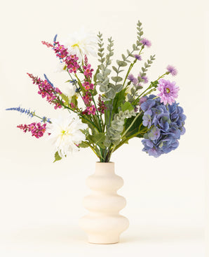 Een crèmekleurige golvende vaas bevat het Zijden kunstbloemen boeket Lavender Kiss, met witte dahlia's, lila kunstbloemen, paarse hortensia's, roze en paarse bloemen en groen gebladerte tegen een effen lichte achtergrond.