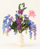 Een witte golvende vaas toont het Zijden kunstbloemen boeket Indigo Dreams - een arrangement van paarse, blauwe en roze kunstbloemen met groene bladeren - tegen een effen lichte achtergrond.