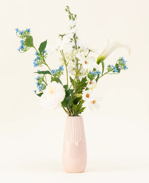 Le vase rose clair contient le bouquet de fleurs artificielles en soie Whispers of White, avec des fleurs blanches, des fleurs bleues et un feuillage vert sur un fond uni et clair.