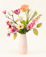 Het Zijden kunstbloemen boeket Spring Awakening, met roze magnolia's, lichtgele klaprozen en oranje en paarse bloemen in een hoge lichtroze keramieken vaas, benadrukt het Nederlands vakmanschap tegen een effen witte achtergrond.