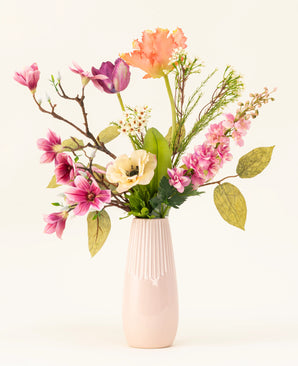 Het Zijden kunstbloemen boeket Spring Awakening, met roze magnolia's, lichtgele klaprozen en oranje en paarse bloemen in een hoge lichtroze keramieken vaas, benadrukt het Nederlands vakmanschap tegen een effen witte achtergrond.