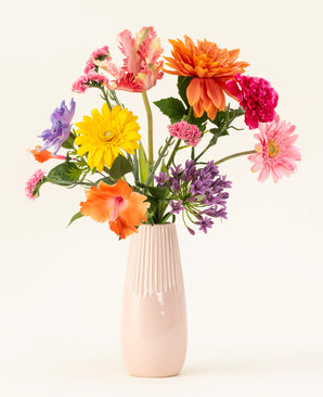 Een lichtroze vaas van keramiek bevat het Zijden kunstbloemen boeket Candy Petals met levendige oranje, gele, roze en paarse zijden bloemen tegen een effen gebroken witte achtergrond.