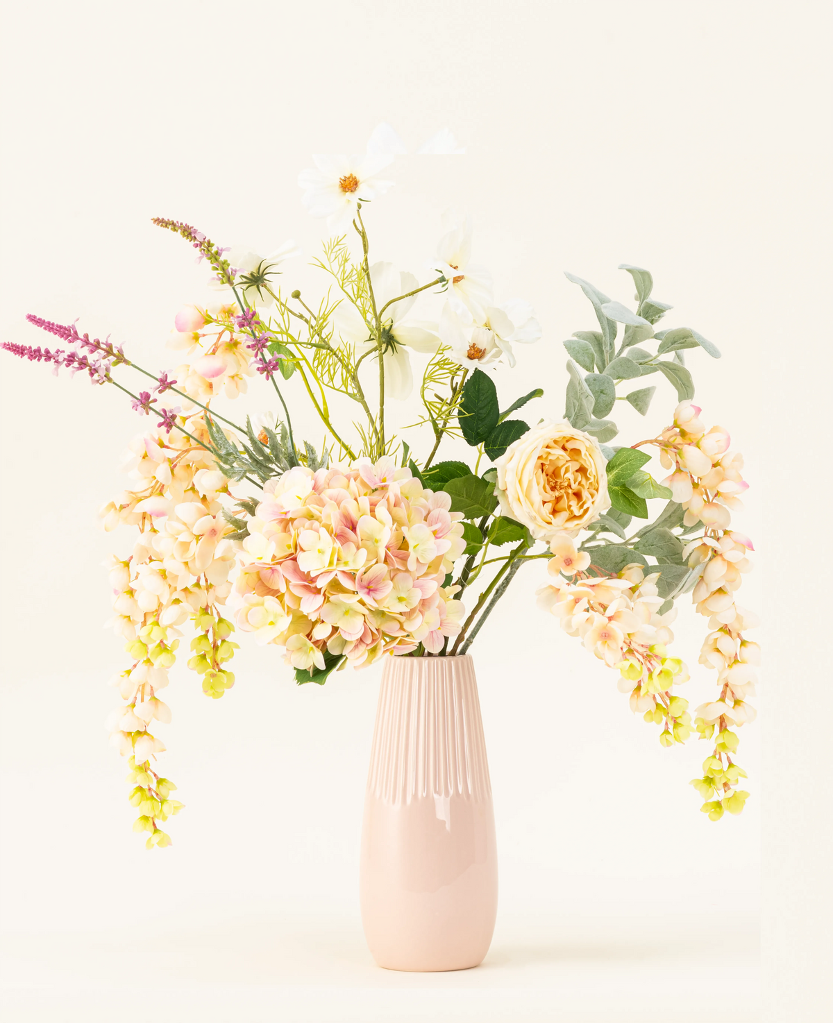 Un vase en céramique rose pastel présente le bouquet de fleurs artificielles en soie Blushing Beauty, composé d'hortensias aux couleurs douces, de roses et de verdure assortie, magnifiquement disposés sur un fond neutre pâle.
